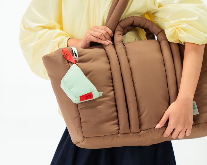 Woman holding a large padded brown tote bag with a pastel Yellow Puft Mini Bag Charm attached to the handle, showcasing Daily Objects tarp-inspired bag collection.